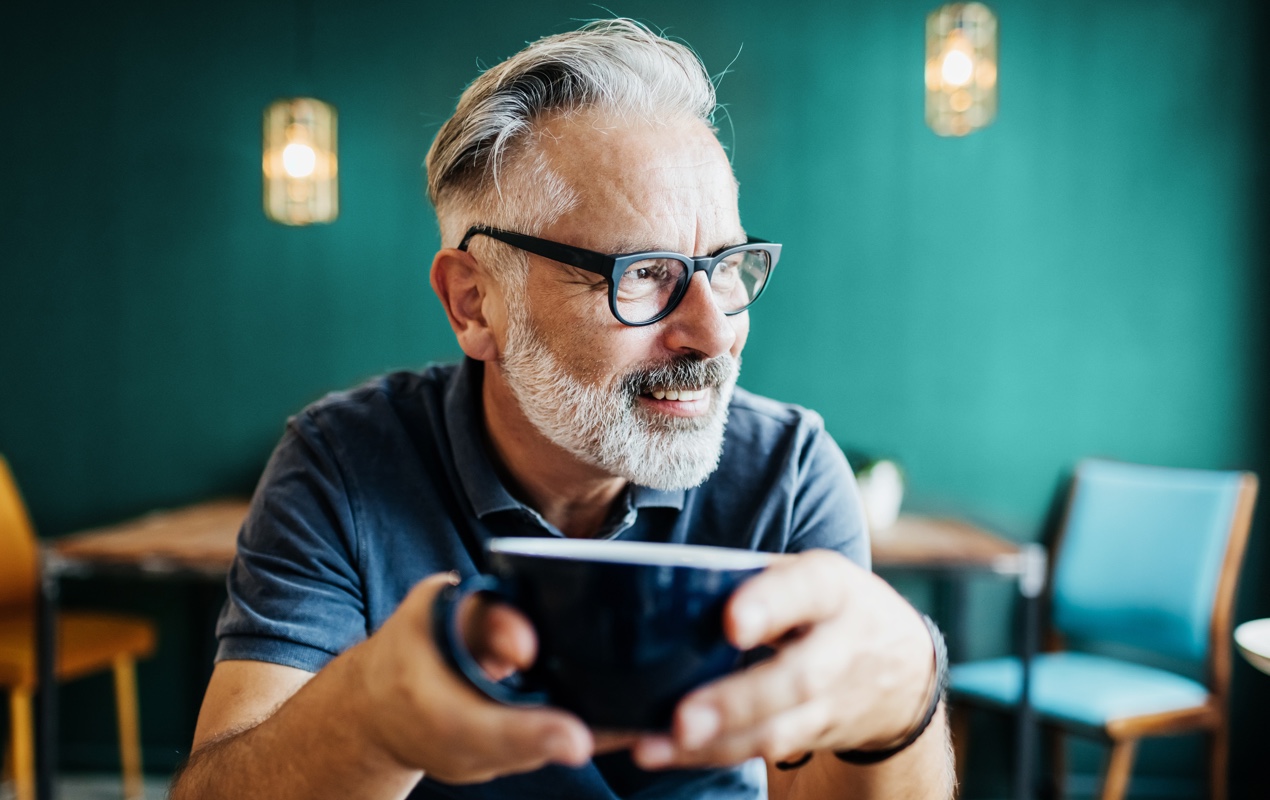 A man is holding a coffee mug and smiling while looking to his left.