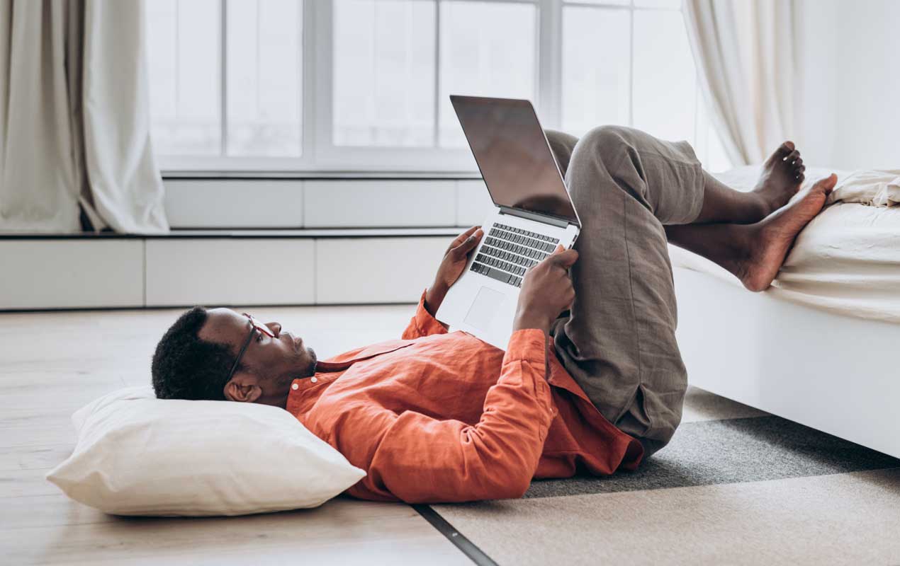 A man is laying on the ground with his feet on the bed and a laptop in his hands.