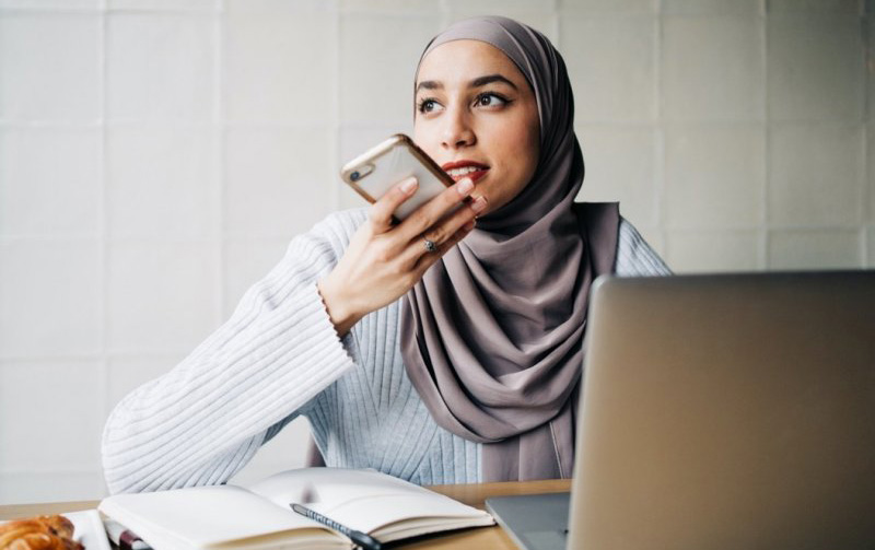 A woman wearing a hijab is speaking on her smartphone while doing research on her laptop.
