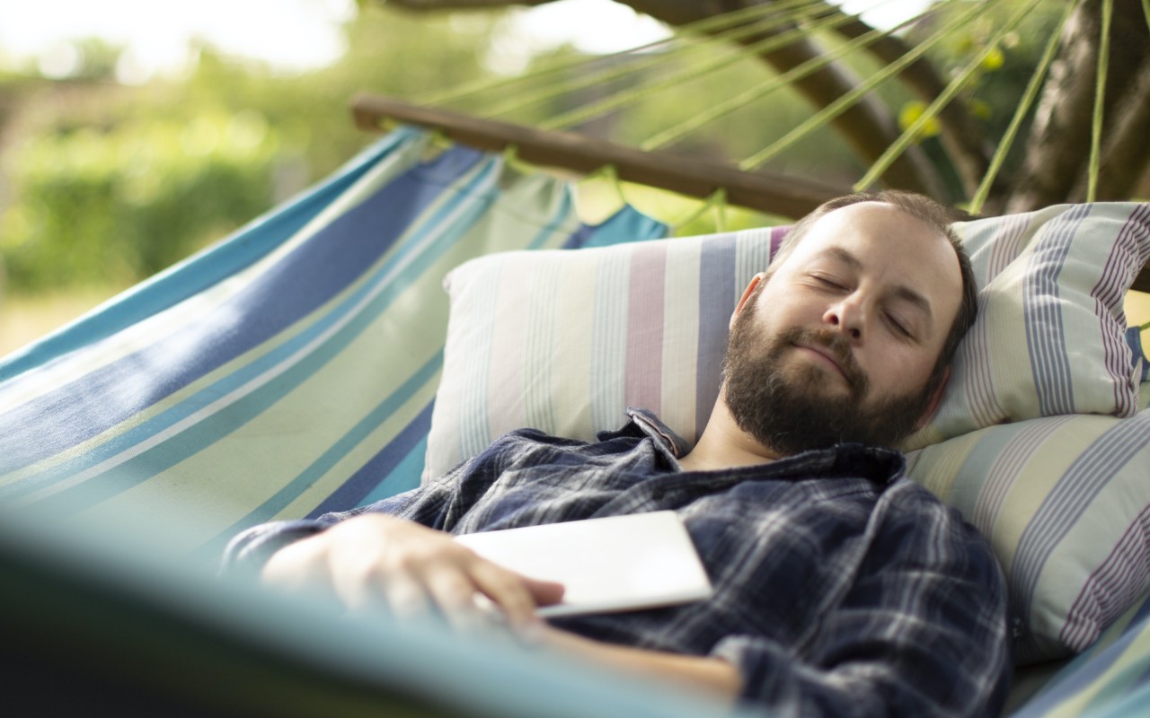 Man in blue plaid flannel shirt, slightly smiling, while relaxing in a hammock outdoors. 