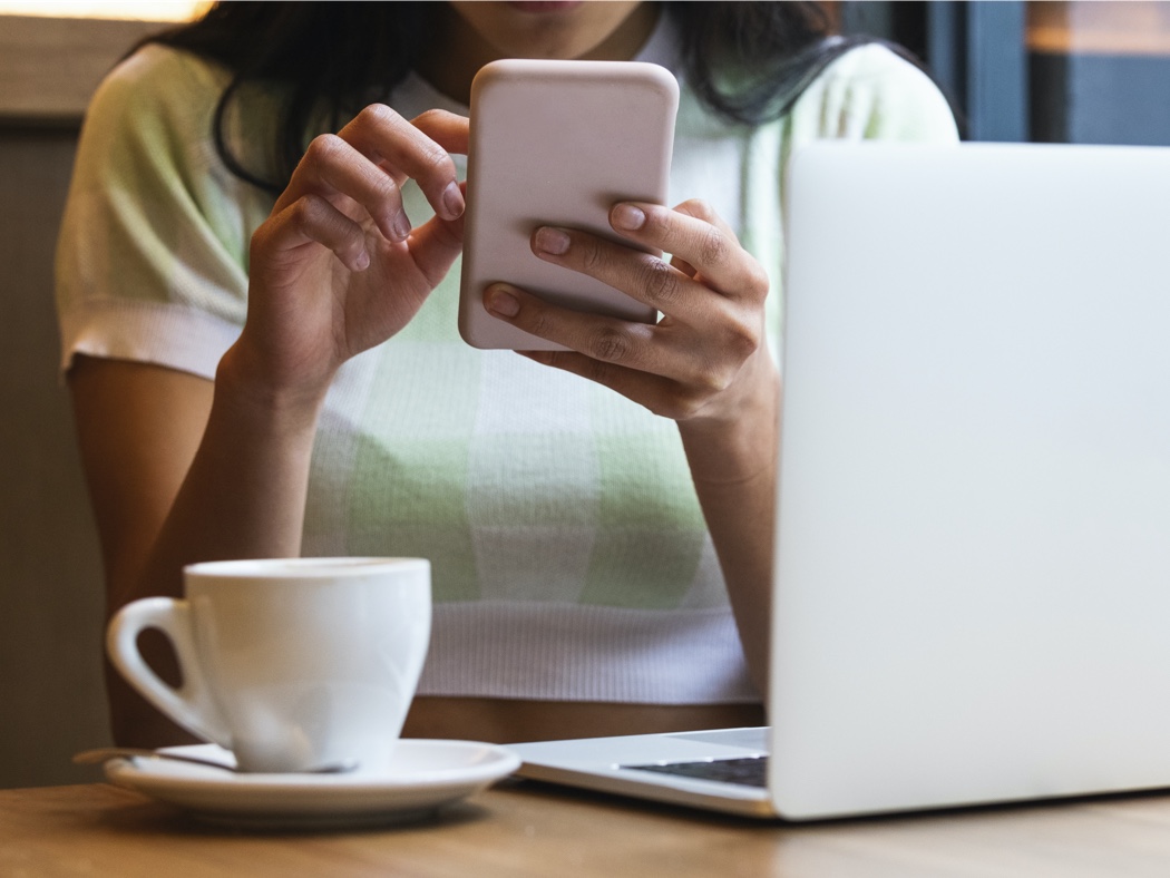 Woman in white and green sweater looking at 403b information on a cell phone, sitting at a table with coffee and laptop. 