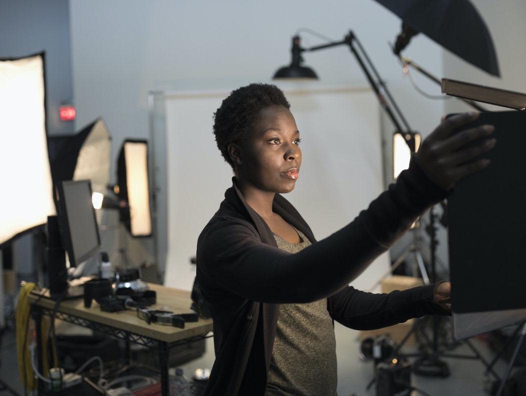 Woman in grey shirt and black sweater in a photo studio adjusting a camera light.