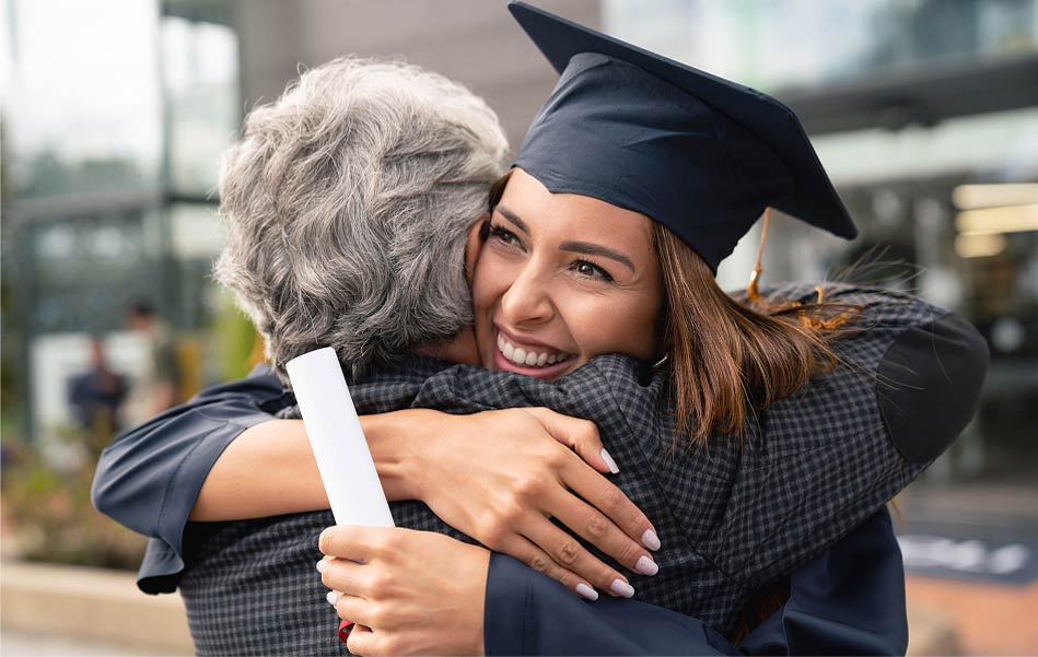 Graduate wearing cap and holding diploma hugging family member.