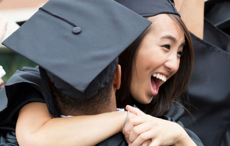 A young man in a graduation cap and gown is hugging a smiling young woman who is also in a graduation cap and gown.