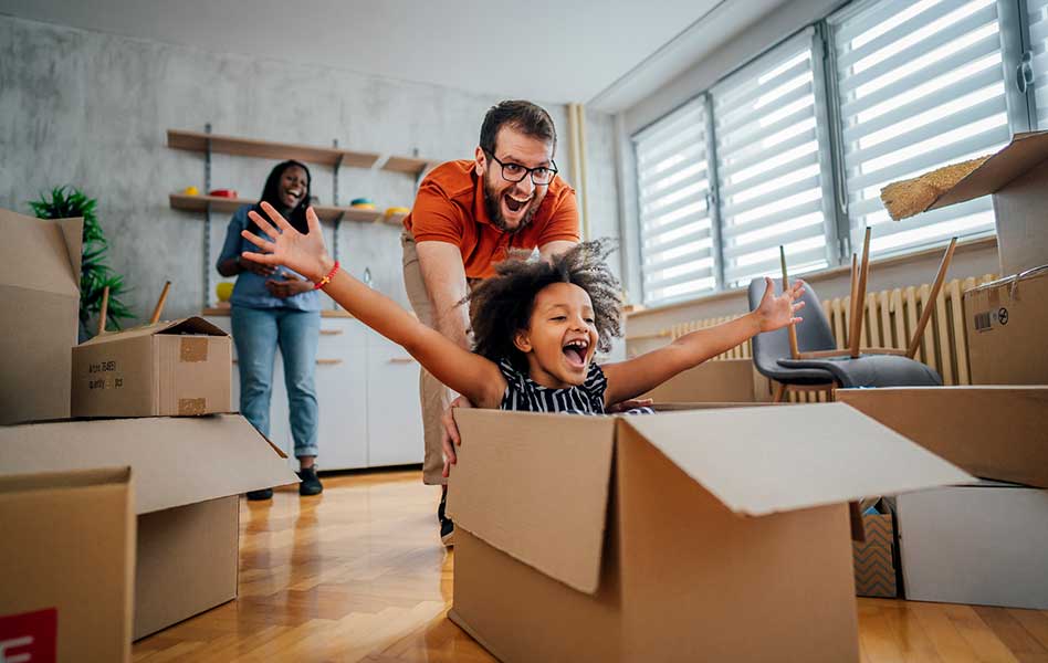 A father is pushing his daughter in a carboard box while she stretches out her arms with a big smile on her face.