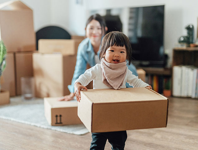 A mother, in the middle of unpacking moving boxes, smiles as her toddler holds a large cardboard box.