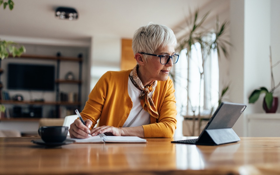 A middle-aged woman is sitting at her dining table learning about Vanguard Roth IRAs from her tablet.