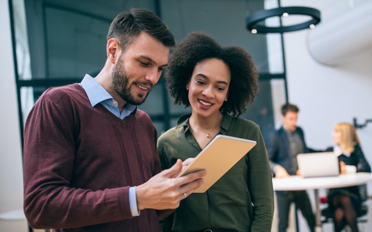 A man and woman standing side-by-side in an office and smiling, looking at a tablet and reviewing organization accounts.