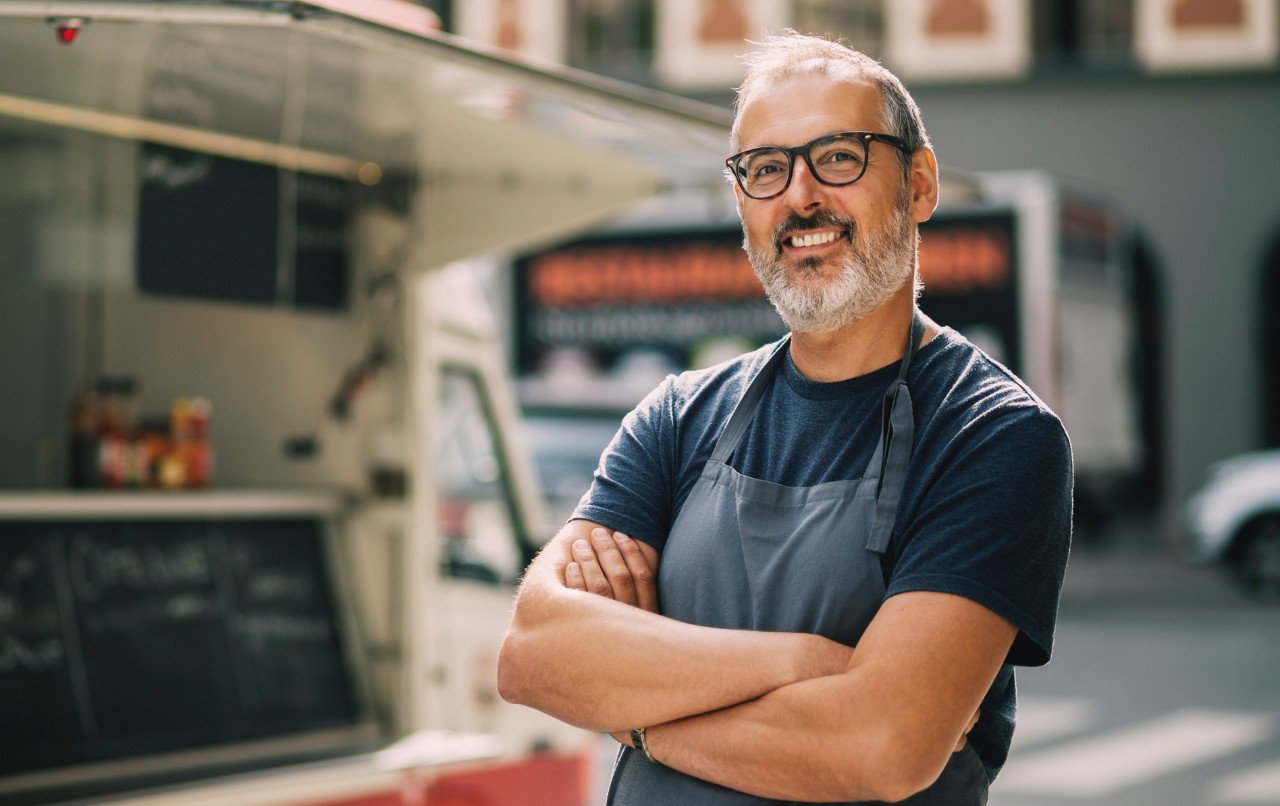 A man wearing, and apron is standing in front of a food truck.
