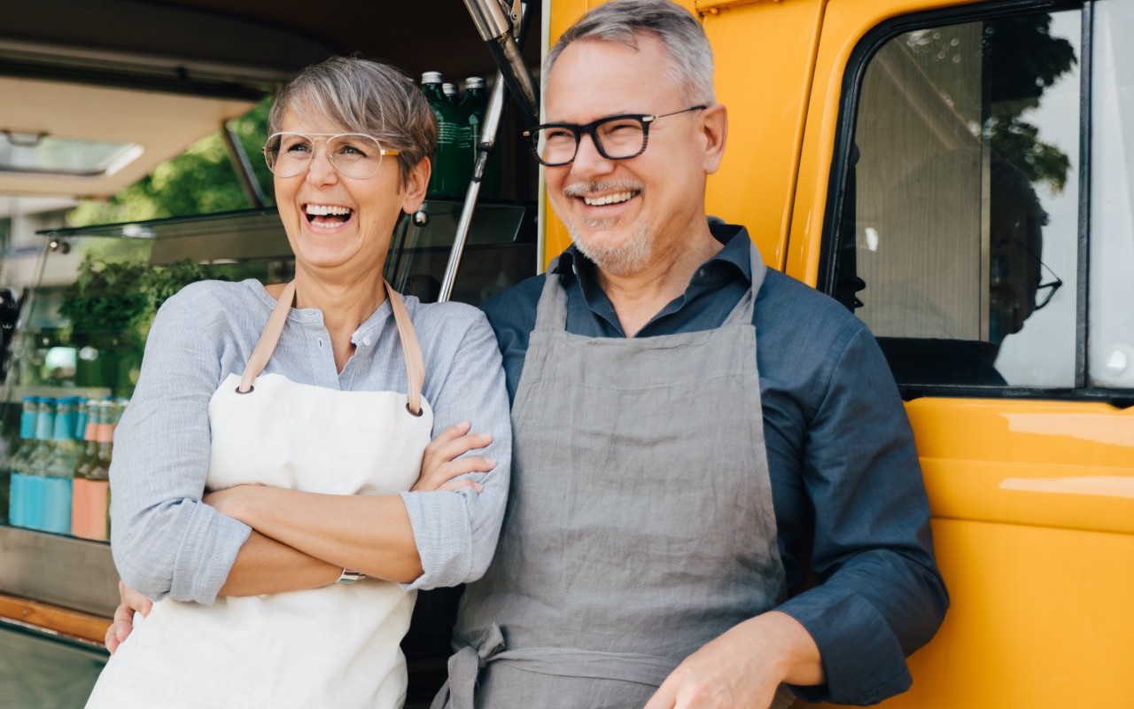 A man and woman, both wearing aprons, laugh together while standing in front of a food truck.