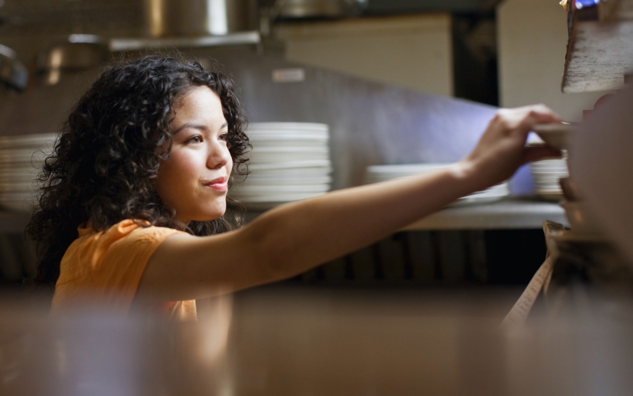A waitress grabbing a plate in a restaurant's kitchen.