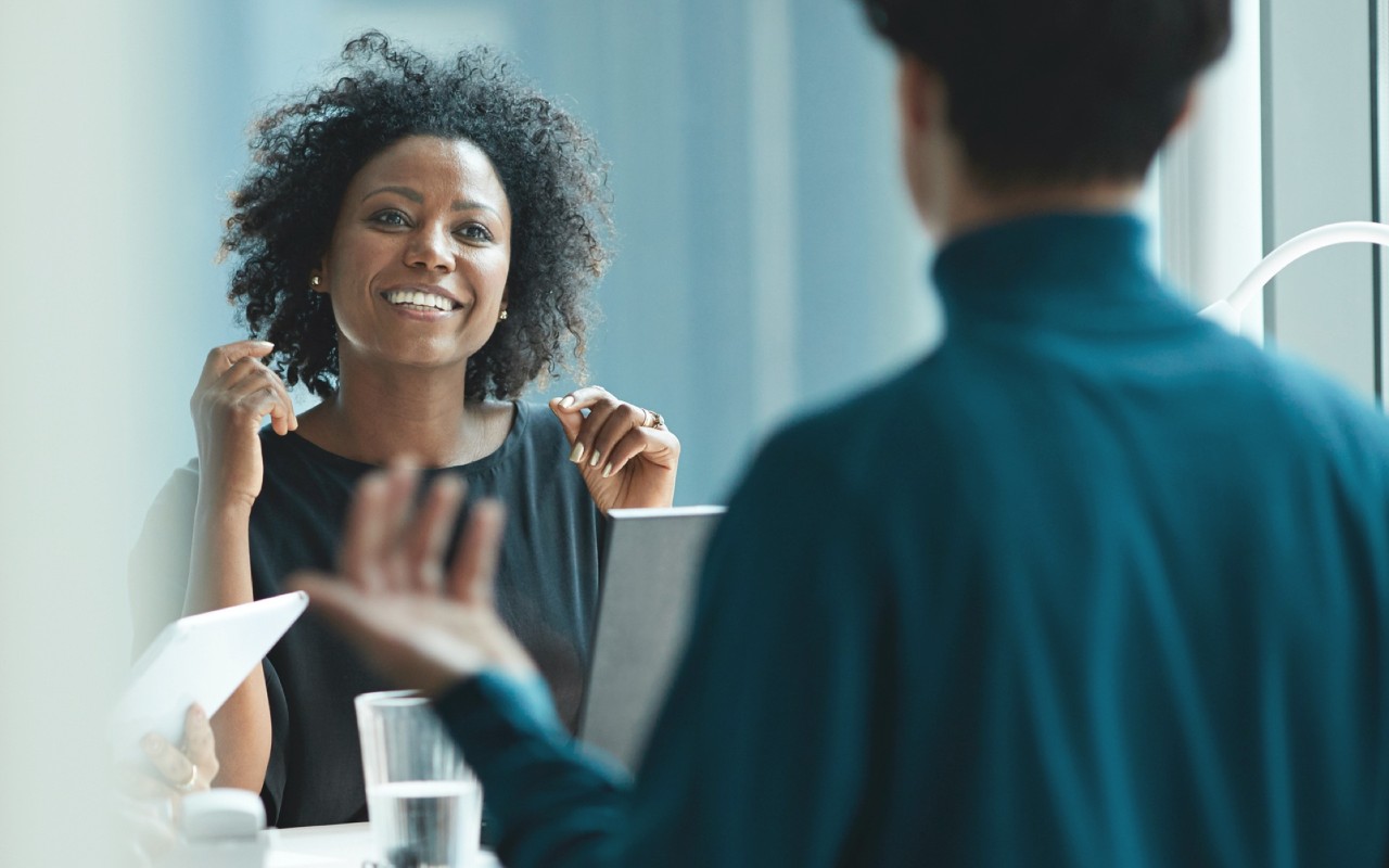 Woman smiling and seated across from a standing person and next to another seated person, talking about Vanguard funds.