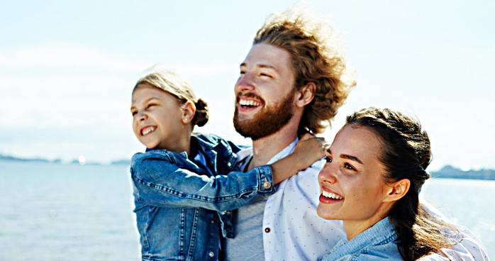 Man holds a young female child with arm around a woman, all smiling, in front of a lake.