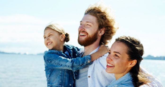 Man holds a young female child with arm around a woman, all smiling, in front of a lake.