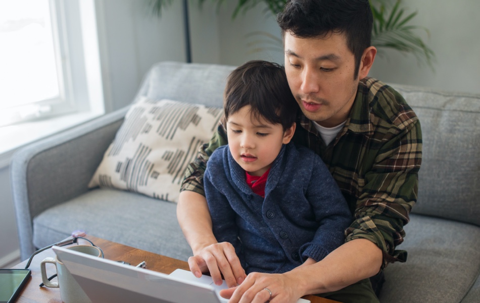 On a couch in a living room, a father and his son view a laptop screen together.