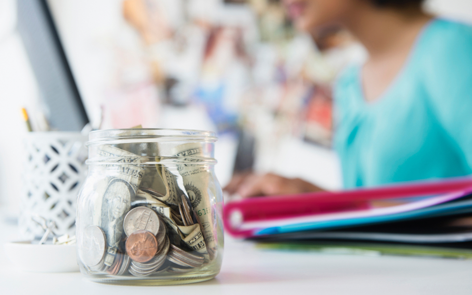 A glass jar of saved change is in focus while a woman who is out of focus is working on her laptop at the same desk.