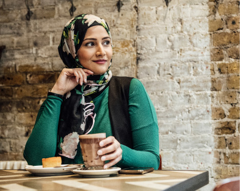 Woman wearing a hijab is sitting at a table with a drink in hand while she look off to her left side.