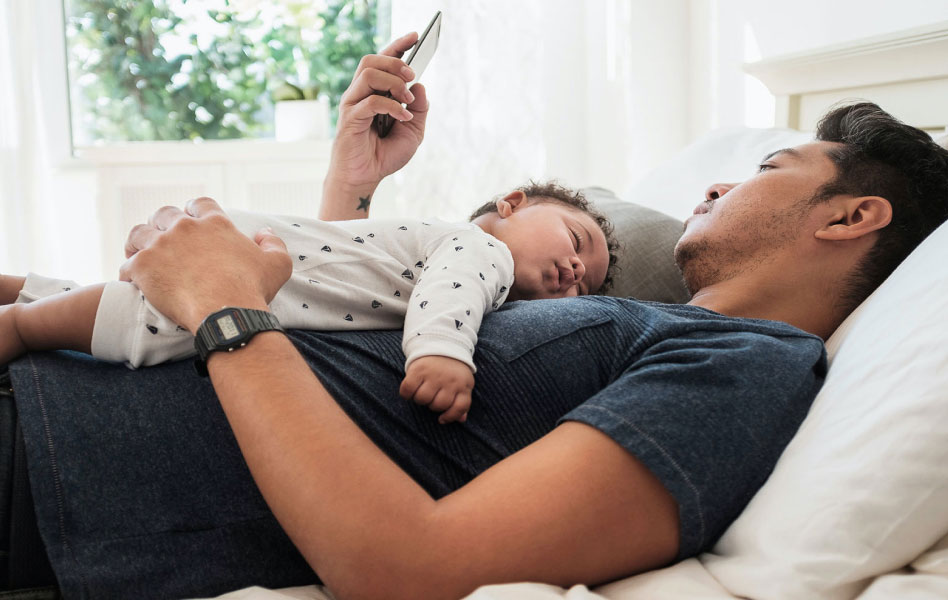 Man with sleeping baby on his chest looks at his phone