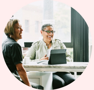 Two women laughing sit in an office with a tablet on the table.