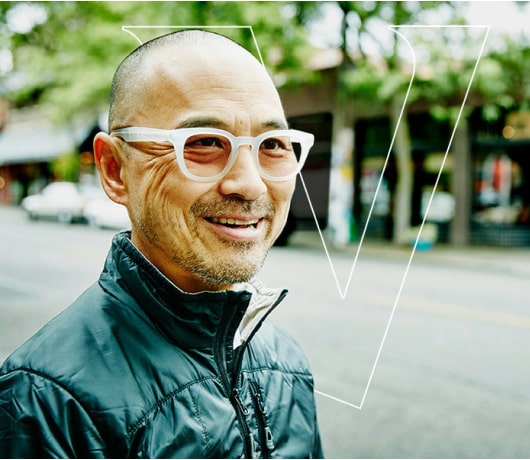 A man with bold, white glasses smiles on a city street. An outline of the Vanguard V is in the background.