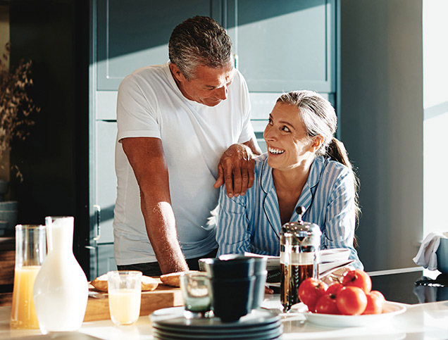 A smiling man and woman are sharing a newspaper in their sunny kitchen..