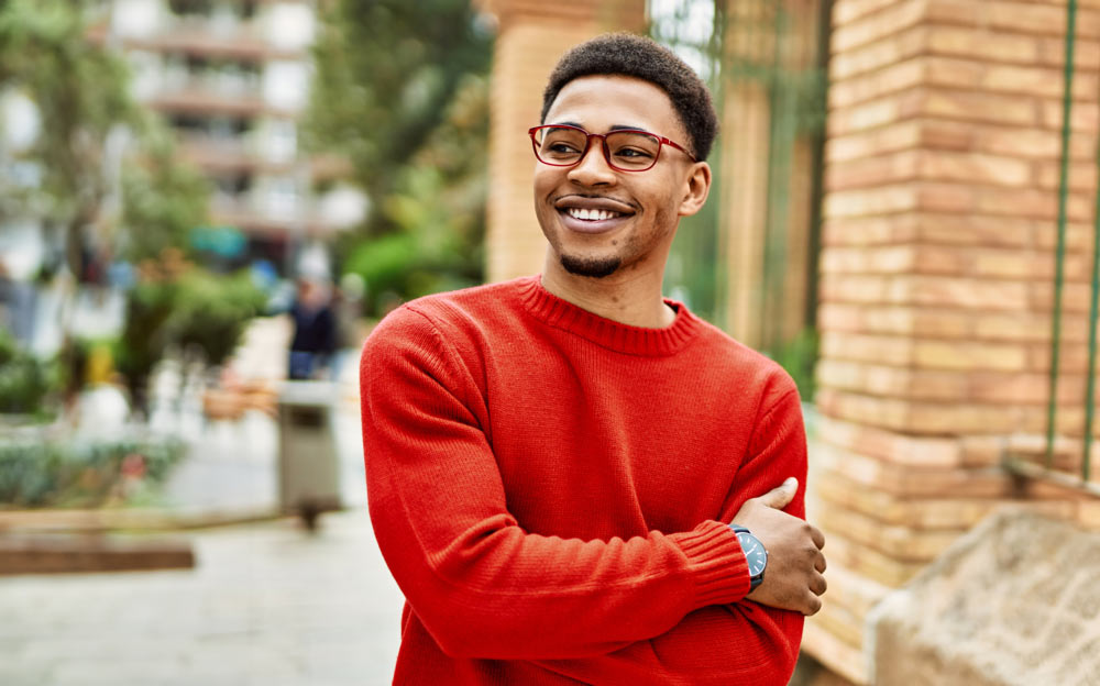 A smiling man in a red sweater stands in a city plaza. 