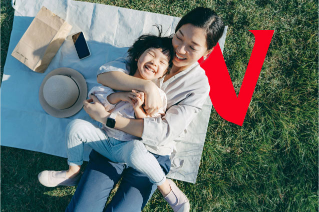 A mother cuddles her laughing child on a picnic blanket outside.