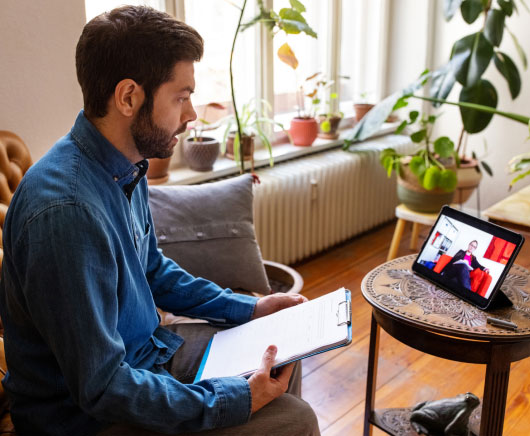 A man holding a clipboard sits in his living room while talking to an advisor on a tablet. 