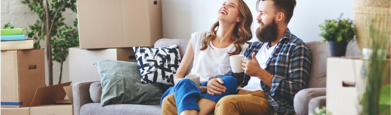 A young couple enjoys a cup of coffee and a laugh while seating on their couch after unpacking boxes in their new house.