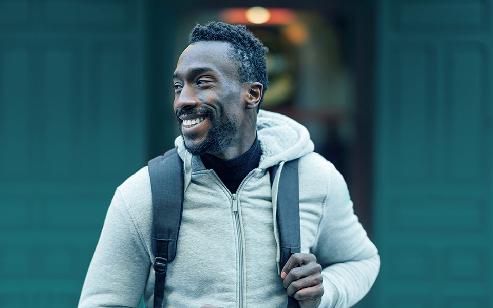 Man with a backpack on a city street, looking to the side and smiling.