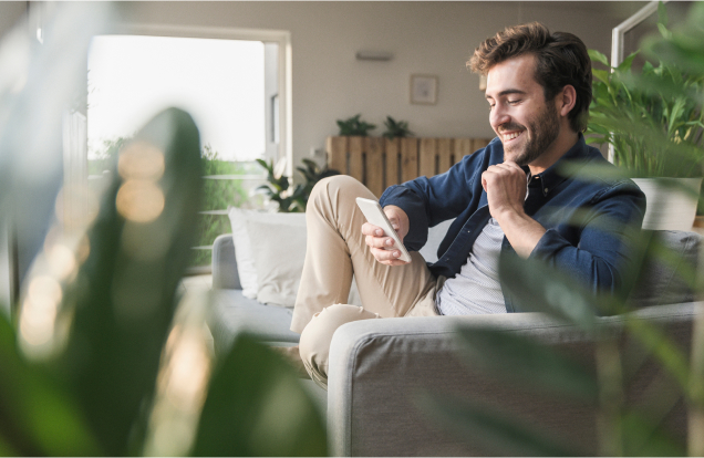 A young man is seated on a couch, smiling at his smartphone screen.