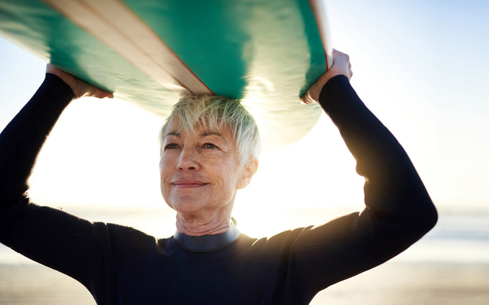 Person in a wetsuit on the beach, looking serene and happy while holding a paddleboard aloft.