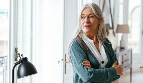 Woman with long white hair looking out of a window with a somber expression.