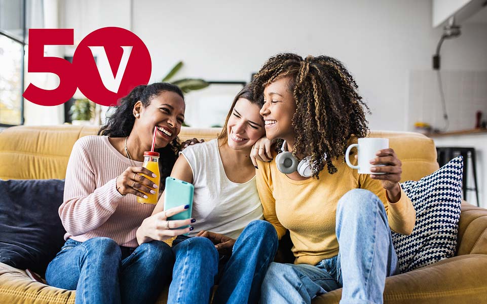 Three friends taking a selfie on the couch together. One women holds a jar of orange juice, one woman holds  a cell phone, and one woman holds a white coffee mug and has headphones around her neck. There's a Vanguard "V" logo, featuring a red number "50" in the background.