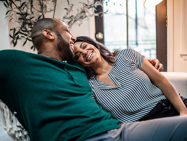 A woman reclines on the couch with her husband, smiling and leaning her head on his shoulder.