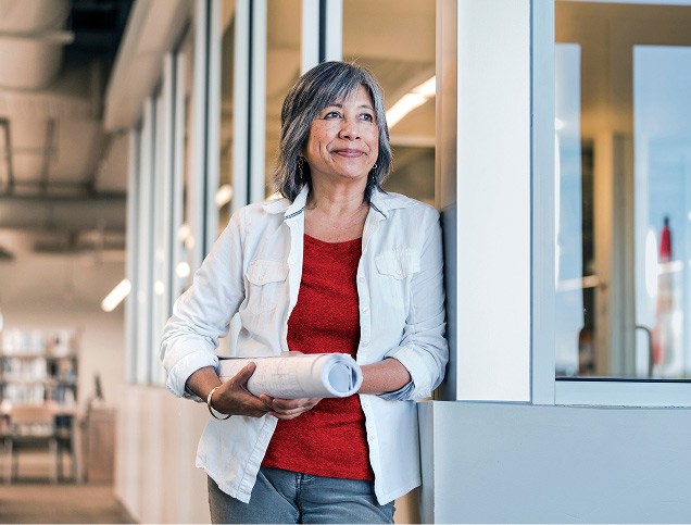 A woman in an office holds a rolled up poster under her arm while leaning against the wall.