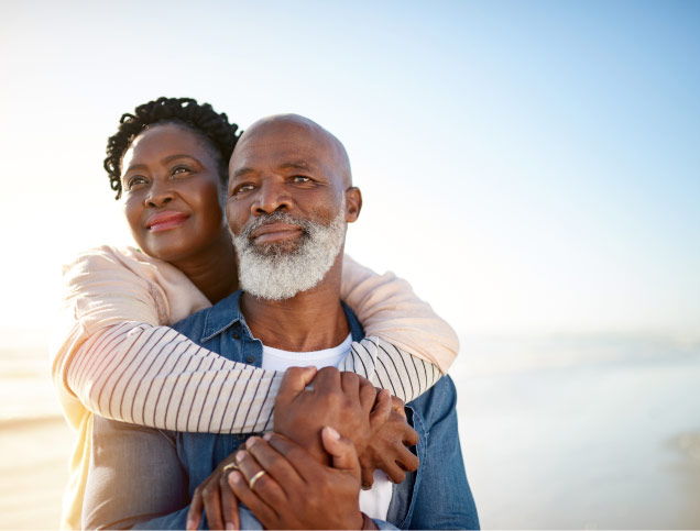 : Older African American couple sharing a tender embrace on a beach, their faces filled with contentment as they gaze into the distance together.