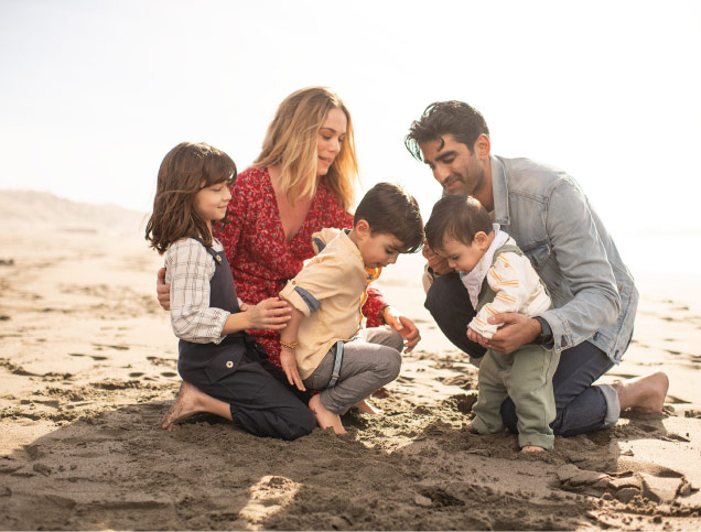 A family of two adults and three young children, all barefoot and smiling at the beach.