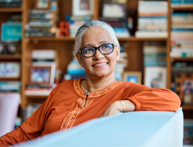 Older adult, wearing glasses and a smile, sitting in a chair before a shelf full of books.
