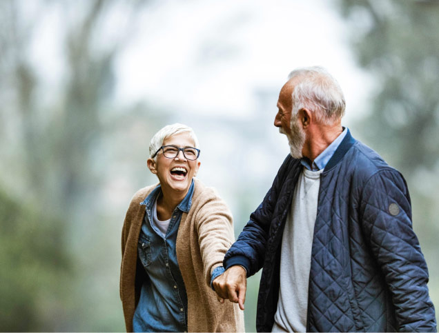 Older adult couple, smiling as they hold hands, while walking down a tree-lined street in the fall.