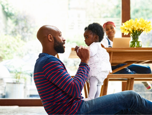 A parent is happily engaging with their baby on the floor. In the background, the other parent sits at a table, attentively watching while working on a laptop.
