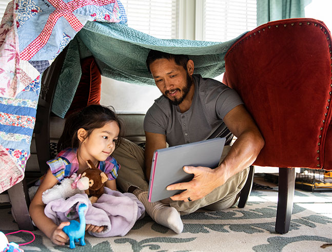 A father reads to his daughter in a fort created out of pillows and blankets.