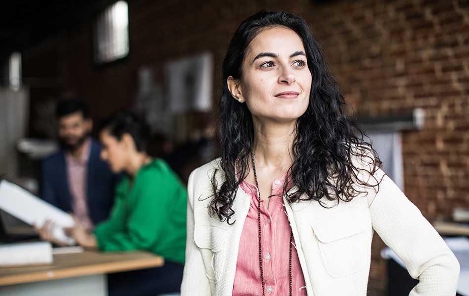 Person stands in an office while others work at a table in the background.