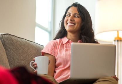 A woman is holding a coffee mug on her lap while using her laptop on a couch.