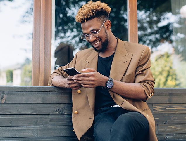 A young man seated on a bench is smiling while looking at his smartphone.