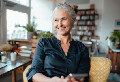 An older woman is holding a smartphone while smiling at someone out of view.