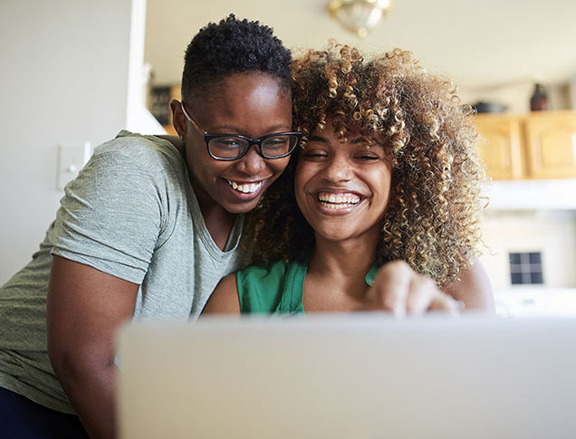 A young woman is smiling and embracing another smiling woman while they both looking into the laptop camera.