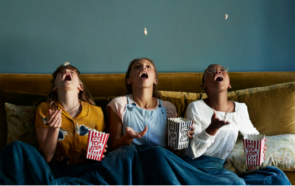 Three children are sitting on a couch and tossing popcorn into their mouths.