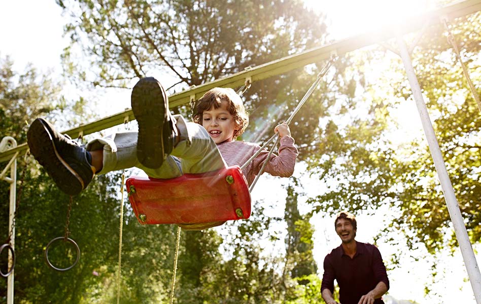 A father is laughing while pushing his smiling son on a swing.