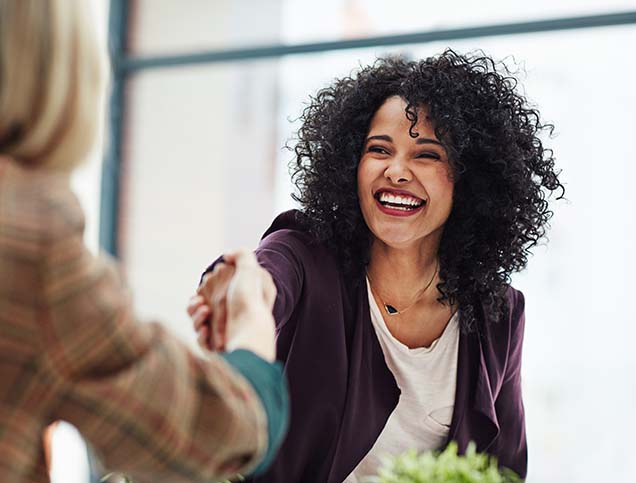 A young woman is smiling brightly while shaking hands with another woman who is out of the frame.